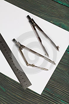 Set for drawing. Two compasses, a metal ruler and a sheet of white paper. They lie on pine boards painted in black and green