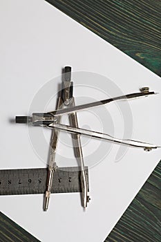 Set for drawing. Two compasses, a metal ruler and a sheet of white paper. They lie on pine boards painted in black and green