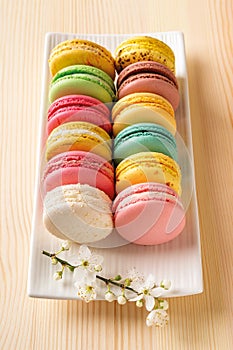 Set of multicolored macaroons and spring cherry blossom flowers on a white rectangular plate against natupal wood table. Sweet