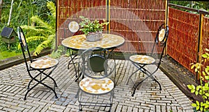 Set of outdoor dining furniture. Mosaic tile and iron chairs and table on a grey brick patio in a lush garden. Cozy