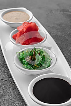 Set of sauces for sushi on a white plate, ginger, soy, sesame, seaweed. dark gray textured background, side view, close-up