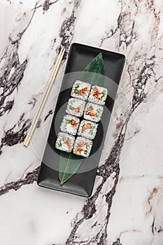 Set of square maki rolls with fried salmon, curd cheese and green bamboo leaf in a black ceramic plate with chopstick on a bright