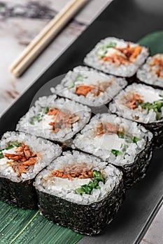 Set of square maki rolls with fried salmon, curd cheese and green bamboo leaf in a black ceramic plate with chopstick on a bright