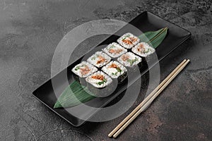 Set of square maki rolls with fried salmon, curd cheese and green bamboo leaf in a black ceramic plate with chopstick on a dark