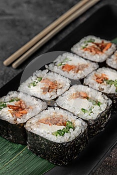 Set of square maki rolls with fried salmon, curd cheese and green bamboo leaf in a black ceramic plate with chopstick on a dark