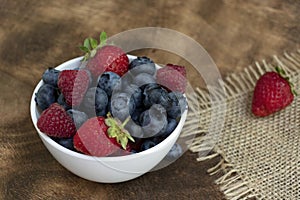 Set of summer berries in a small white bowl on a napkin and wooden background. Blueberries, raspberries, and strawberries are a