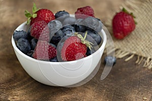 Set of summer berries in a small white bowl on a napkin and wooden background. Blueberries, raspberries, and strawberries are a