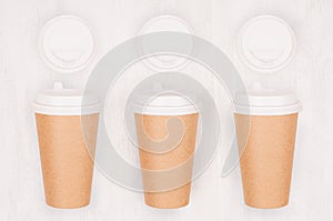 Set of three coffee kraft paper cups and blank cap on light wood board, top view.