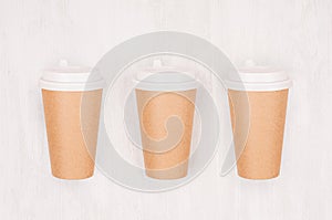 Set of three coffee kraft paper cups with cap on light wood board, top view.