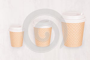 Set of three different sizes ribbed coffee kraft paper cups with cap on light wood board, top view.