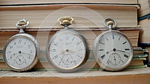 Set of three silver antique pocket watch with against background of stacks of books. Retro clock near old textbooks with