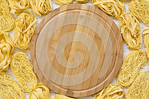 Set of various uncooked pasta with bamboo board in the center on white wooden background, top view.