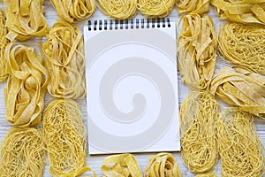 Set of various uncooked pasta with blank notepad on white wooden background