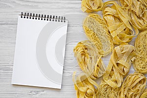 Set of various uncooked pasta with notebook on a white wooden background, flat lay.