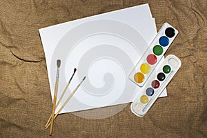 Set of watercolor paints, brushes for painting and blank white paper sheet of sketchbook.