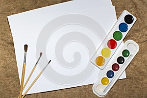Set of watercolor paints, brushes for painting and blank white paper sheet of sketchbook.