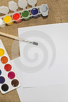 Set of watercolor paints, brushes for painting and blank white paper sheet of sketchbook.