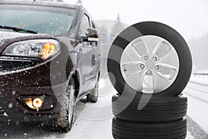 Set of winter tires near broken car with open trunk on forest highway during snowfall.
