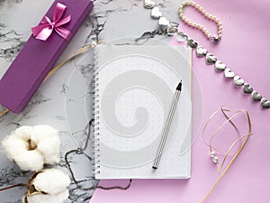 Set of women`s accessories desk - notebook with pen, gifts, jewelry, bracelet, cotton flowers on pink marble background. Workspac