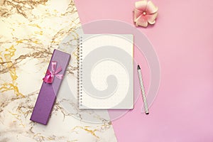 Set of women`s accessories desk - notebook with pen, pink box gifts, flower on pink marble background. Workspace table for beauty