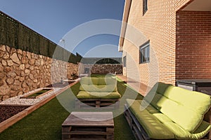 Settee of wooden pallet sofas with green cushions, matching coffee table next to a brick house with a garden and artificial grass