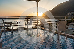 The setting sun illuminates the summer terrace with cozy tables and chairs against the backdrop of the sea and mountains. Travel