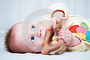 A seven-month-old girl with blue eyes lies on her side in a bright room and nibbles on a wooden toy, looking up. The child`s teet