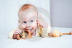 A seven-month-old girl with blue eyes lies on her stomach in a bright room and nibbles on a wooden toy, looking up. The child`s