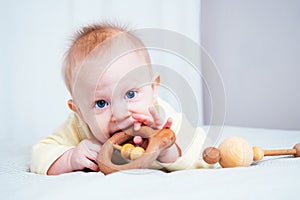 A seven-month-old girl with blue eyes lies on her stomach in a bright room and nibbles a wooden toy, looks at the camera the child