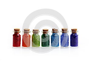 Seven tiny glass bottles with a cork stopper, filled with a rainbow colours of beads, on a white background