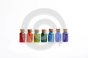 Seven tiny glass bottles with a cork stopper, filled with a rainbow colours of beads, on a white background