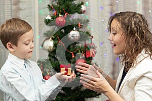 A seven-year-old boy gives his mother a gift in a box for the New Year against the backdrop of a Christmas tree at home