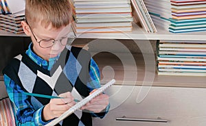 Seven-year-old boy with glasses thinking and draws something in a sketchbook sitting among the books.