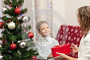 A seven-year-old boy in a smart shirt gives his mother a gift in a box for the New Year against the backdrop of a Christmas tree a