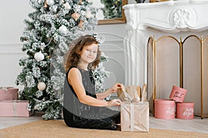 A seven-year-old girl in a black dress with sequins sits near the fireplace at the Christmas tree and opens a New Year`s gift