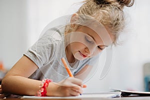 A seven-year-old girl sits at home at a table and writes in a notebook, completing a learning task or repeating lessons.