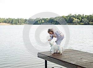 A seven-year-old girl with a small white dog stands on a bridge near the lake and looks at the water.