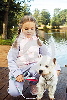 A seven-year-old girl with a small white dog stands on a bridge near the lake and looks at the water.