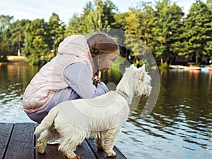 A seven-year-old girl with a small white dog stands on a bridge near the lake and looks at the water.