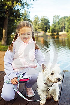 A seven-year-old girl with a small white dog stands on a bridge near the lake and looks at the water.