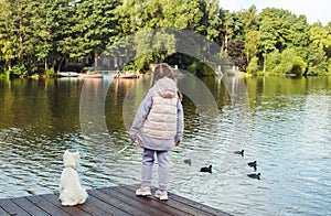 A seven-year-old girl with a small white dog stands on a bridge near the lake and looks at the water.
