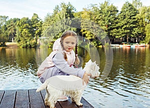 A seven-year-old girl with a small white dog stands on a bridge near the lake and looks at the water.