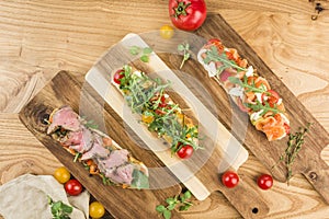 Several bruschetta with salmon, roast beef and mozzarella with cherry tomatoes on a board on a light wooden background top view