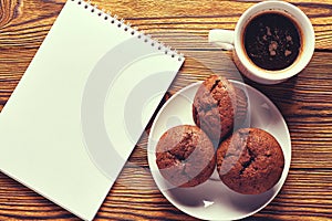 Several dark chocolate dough muffins with a cup of coffee and a notepad on a wooden table