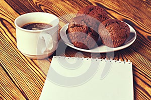 Several dark chocolate dough muffins with a cup of coffee and a notepad on a wooden table