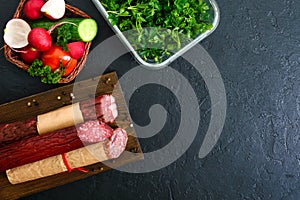 Several kinds of sausage, fresh vegetables and greens on a black background.