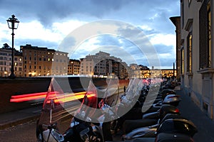 Several motorbikes parked in a row along the street with light trails of a car and cityscape of Florence