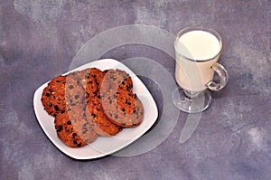 Several oatmeal chocolate chip cookies on a plate and a glass of hot milk on a gray abstract background