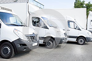 Several white row commercial delivery vans and service van, trucks and car in front of factory warehouse