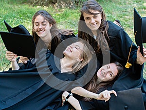 Several young smiling female students in black graduation gowns lie on the grass in the university park